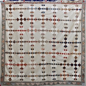 May include: A large, cream-colored quilt with a repeating pattern of squares and diamonds. The quilt features a grid-like design with alternating light and darker squares, and a patterned border. The color palette includes cream, brown, red, and blue.