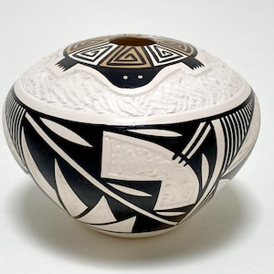 May include: A white ceramic pot with black and brown geometric designs. The pot features a carved turtle design on the top.