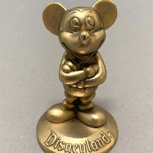 Op de afbeelding: Een gouden Mickey Mouse-figuur dat op een voet staat met het woord "Disneyland" erop.