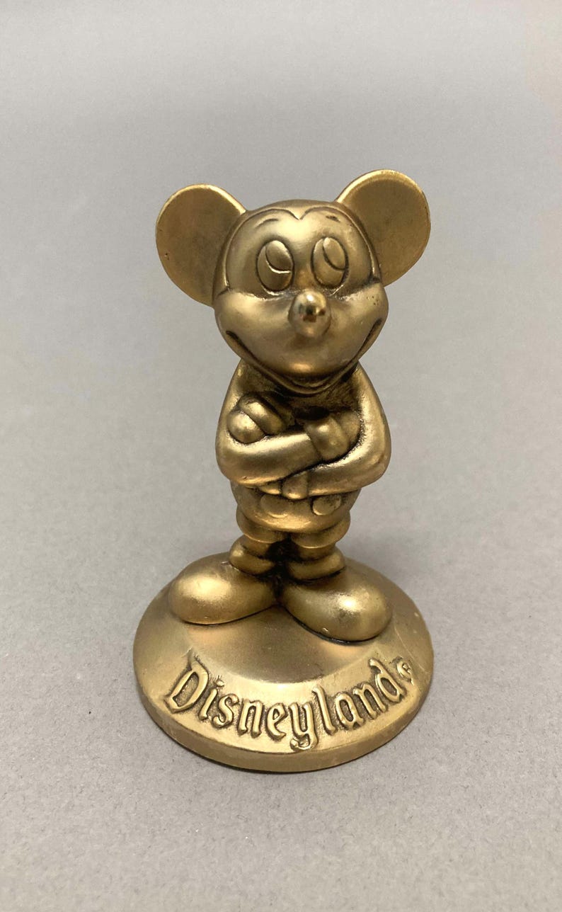 Op de afbeelding: Een gouden Mickey Mouse-figuur dat op een voet staat met het woord "Disneyland" erop.