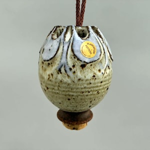 May include: A ceramic hanging ornament with a speckled cream and blue glaze, featuring a scalloped top and a brown cord. A circular gold sticker reads Plantation Potters Plantation, FL. The base has a dark brown flared rim and a cork stopper.