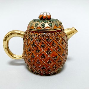 May include: A small, gold-colored teapot with a lid decorated with green and white enamel. The teapot is shaped like a pineapple and has a detailed pattern of gold and orange enamel.