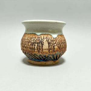 May include: A small ceramic pot with a flared rim, featuring a smooth white glazed upper section that drips over a textured, crackled brown body. The base displays a dark blue, carved, repeating pattern around the bottom circumference.