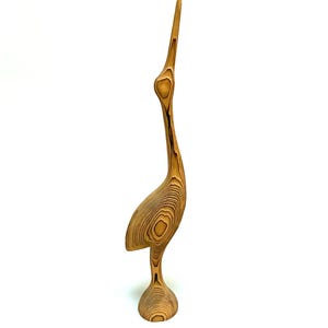 May include: A tall, slender wooden sculpture of a bird with a long, pointed beak and neck. The object features a prominent, swirling natural wood grain pattern in shades of light tan and dark brown, standing on a circular, flared base.
