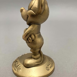 Op de afbeelding: Een gouden beeldje van Mickey Mouse dat op een ronde basis staat. De basis heeft de tekst "Walt Disney Productions" erop.