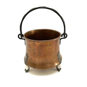 May include: A copper cauldron with a black wrought iron handle and decorative feet. The cauldron has a round body and a dark interior. The handle is arched and attached to the cauldron with decorative brackets.