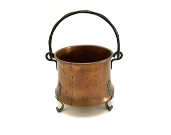 May include: A copper cauldron with a black wrought iron handle and decorative feet. The cauldron has a round body and a dark interior. The handle is arched and attached to the cauldron with decorative brackets.