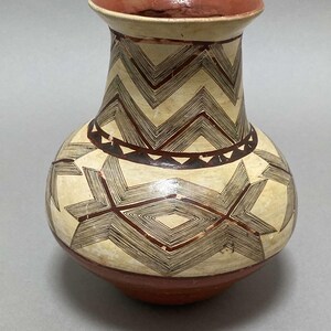 May include: A ceramic vase with a cream-colored body and a geometric pattern in brown and black. The vase has a rounded base and a narrow neck.