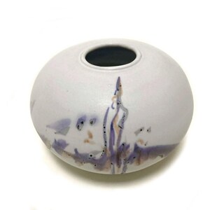 May include: A small, round, white ceramic vase with a dark interior. The vase features a painted design in shades of purple, orange, and brown. The design appears to be an abstract landscape or floral motif.