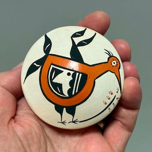 May include: A circular ceramic object with a cream-colored base featuring a stylized orange bird design. The bird has black wings, a black tail, and a black and white patterned body. Small orange dots trail behind the bird's long, pointed beak.