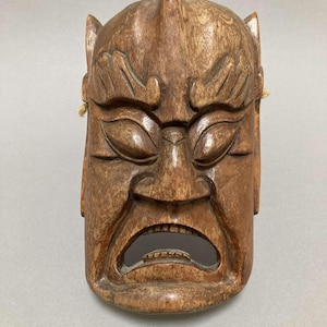 May include: A wooden mask with a carved face, featuring a wide open mouth and a single horn on the forehead. The mask is brown and has a distressed finish.