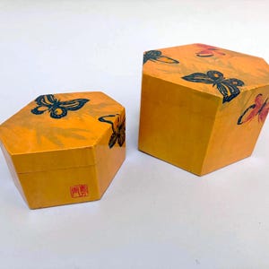 May include: Two hexagonal boxes in a vibrant yellow finish, each featuring black and red butterfly illustrations on the lids and sides. The smaller box displays a red square stamp with Asian characters on its front face.