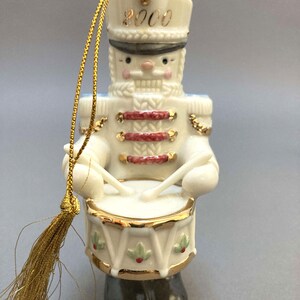 May include: A ceramic Christmas ornament shaped like a toy soldier drummer. The ornament is white with gold accents, red details, and black boots. The year "2000" is printed on the hat. A gold tassel and string are attached for hanging.