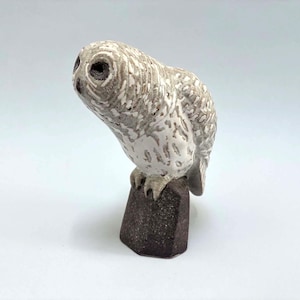 May include: A ceramic owl figurine featuring a textured white and gray plumage pattern. The owl is perched on a dark brown, faceted, hexagonal base. The bird is depicted with its head tilted slightly to the side, showcasing detailed dark eyes.