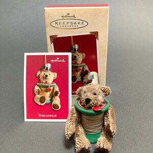 May include: A brown teddy bear ornament sitting on a spool of thread. The bear is wearing a thimble on its head and a red heart on its chest. The ornament is labeled "Threadbear" and is a Hallmark Keepsake Ornament.