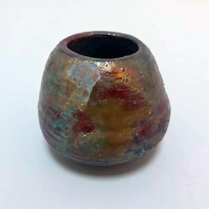 May include: A small, rounded ceramic vessel with a wide opening and a textured, multi-colored surface. The exterior features a mottled blend of metallic gold, deep red, and oxidized blue-green tones, creating a weathered, rustic appearance.