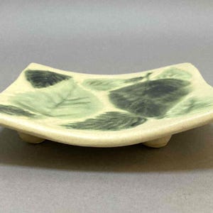May include: A square ceramic dish with a leaf design. The dish is off-white with green and dark green leaf patterns. It has a slightly curved shape and rests on three small feet.
