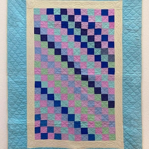 May include: A handmade quilt with a light blue border and a central patchwork design. The patchwork features a grid of squares in shades of blue, pink, and green. The quilt has a cream-colored inner border and a diamond-patterned stitching.