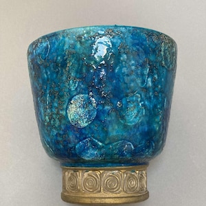 May include: A turquoise ceramic bowl with a textured surface and a gold-colored base. The bowl has a unique, mottled appearance with darker blue and silver accents. The base features a repeating spiral design. The bowl is approximately 5 inches tall.