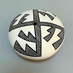 May include: A round, white ceramic vessel with a flat top featuring a black geometric pattern. The design consists of four symmetrical, angular shapes radiating from a small, centered circular opening, all filled with fine, parallel black lines.