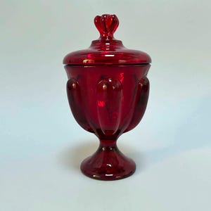 May include: A deep red glass candy dish with a matching lid. The vessel features a pedestal base and a series of vertical, rounded indentations around the bowl. The lid is topped with a small, decorative finial handle, creating a cohesive look.