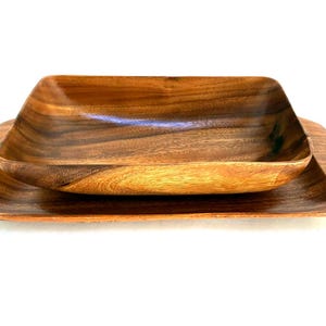 May include: A set of two wooden serving dishes. The top dish is a square bowl with rounded corners, and the bottom dish is a square tray. Both are made of wood with a natural grain and a warm brown color.