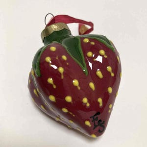 May include: A ceramic strawberry ornament with a glossy finish. The ornament is primarily red with yellow speckles, green leaves, and a gold-colored top. A red ribbon is attached for hanging. The ornament has a signature in black.