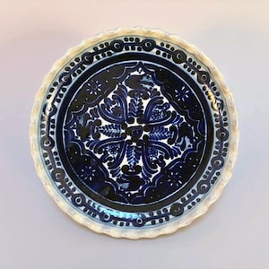 May include: A round ceramic plate with a scalloped edge featuring a deep blue and white hand-painted floral pattern. The center displays a symmetrical botanical design surrounded by a circular border of dots and lines on a white background.