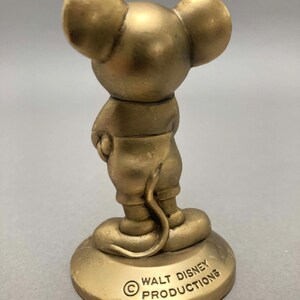 Op de afbeelding: Een gouden beeldje van Mickey Mouse dat op een ronde basis staat. De basis heeft de tekst "&copy; WALT DISNEY PRODUCTIONS" erop gegraveerd.