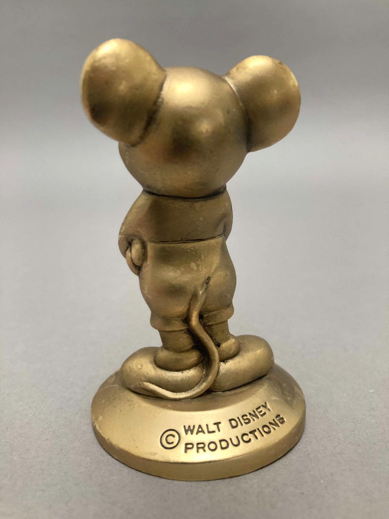 Op de afbeelding: Een gouden beeldje van Mickey Mouse dat op een ronde basis staat. De basis heeft de tekst "&copy; WALT DISNEY PRODUCTIONS" erop gegraveerd.