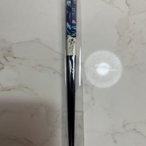 May include: A pair of black chopsticks in a clear plastic sleeve. The top portion of the chopsticks features a blue and white wave design with a mountain scene. The chopsticks are a traditional Asian dining utensil.