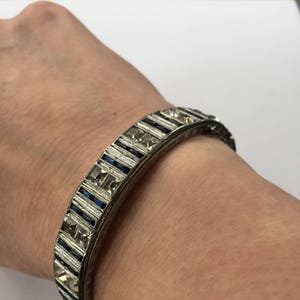 May include: A silver bangle bracelet with a row of clear and blue square-shaped stones set in a geometric pattern.
