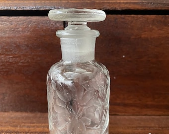May include: A clear glass bottle with a textured, floral design and a simple glass stopper. The bottle is sitting on a wooden surface.