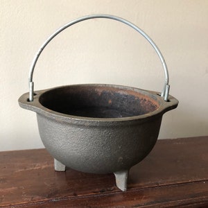 May include: A dark gray cast iron cauldron with three short legs and a curved metal handle. The cauldron has a textured surface and a wide opening. The interior shows signs of use.