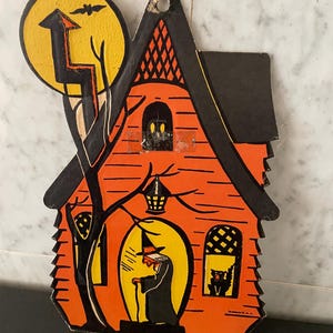 May include: A die-cut cardboard Halloween decoration featuring an orange house with a black roof, a yellow moon with a bat, a witch standing in the doorway, a black cat in a window, and a bare tree with a lightning bolt shape against the moon.
