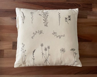 May include: A white pillow with black line drawings of various flowers and plants. The pillow is made of a soft, natural fabric.