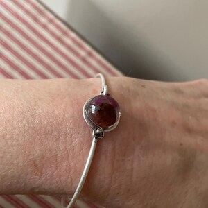 May include: A silver bangle bracelet with a large, round, reddish-brown stone at the center. The stone is set in a silver bezel, and the bracelet has a simple, elegant design. The bracelet is shown on a wrist.