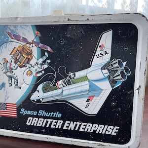 May include: Vintage metal lunchbox featuring a space shuttle, astronauts, and a satellite against a starry background. The lunchbox has the words "Space Shuttle Orbiter Enterprise" and the American flag.