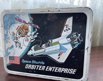 May include: Vintage metal lunchbox featuring a space shuttle, astronauts, and a satellite against a starry background. The lunchbox has the words "Space Shuttle Orbiter Enterprise" and the American flag.