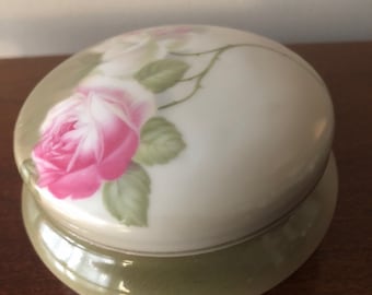 May include: A white porcelain trinket box with a green base. The lid features a hand-painted design of pink and white roses with green leaves.