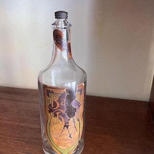 May include: Clear glass perfume bottle with a dark metal cap. The label is a shield shape with the name "Bar R" and "The Bonheur Perfume" printed on it. The label has a brown, orange, and purple color scheme.