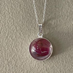 May include: A silver-toned necklace featuring a circular pendant with a smooth, clear domed surface. Inside the pendant, there are deep magenta and dark red organic-looking inclusions set against a lighter, translucent background.