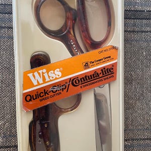 May include: A pair of Wiss Quick-Clip Contura-lite dressmakers shears, 8 inches long, in a clear plastic package. The scissors have brown tortoise shell handles and a silver blade. The orange label reads "Quick-Clip" and "Contura-lite".