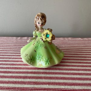 May include: A porcelain figurine of a girl in a green dress holding a yellow flower with a green gem in the centre. The figurine is on a red and white striped surface.
