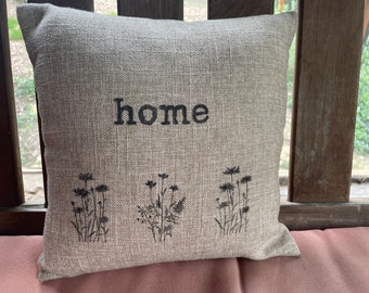 May include: A beige linen pillow with the word "home" embroidered in black. The pillow has three black embroidered flower designs at the bottom.