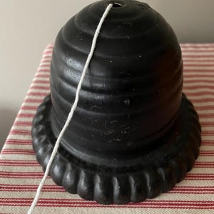 May include: A black, decorative, cast iron lamp shade holder with a scalloped base. A white string extends from the top of the holder. The holder sits on a red and white striped fabric.