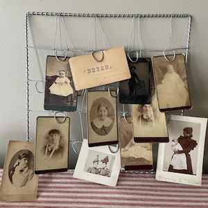 May include: A collection of antique photographs displayed on a wire grid. The sepia-toned images depict portraits of people, including babies and adults. The photos are clipped to the grid with metal clips, creating a vintage aesthetic.
