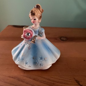 May include: A porcelain figurine of a girl in a blue dress holding a pink rose with a rhinestone centre. The girl has blonde hair and is wearing a gold crown.