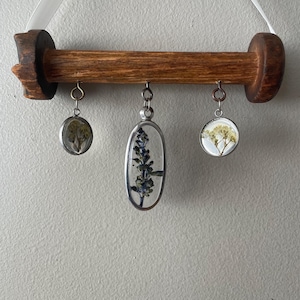 May include: A wooden dowel rod with three hanging glass ornaments. Each ornament contains pressed flowers and is suspended from a silver chain. The dowel rod is attached to a white ribbon.