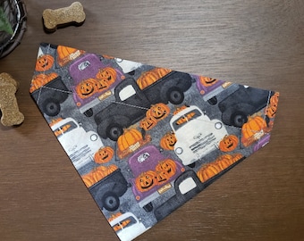 May include: A triangular pet bandana featuring a dark gray background with a repeating pattern of vintage pickup trucks in black, white, and purple. The trucks are loaded with orange jack-o'-lantern pumpkins, creating a festive seasonal design.